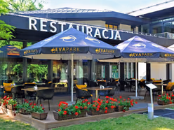 Taras restauracji pod granatowymi parasolami z logo EVA Park, skrzynki z czerwonymi pelargoniami