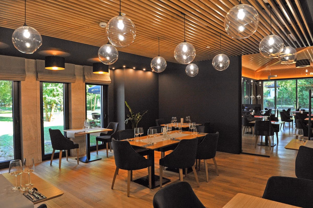 Wnętrze restauracji z drewnianym sufitem listwowym i szklanymi kulami lamp, przez przeszklenie widać zieleń Parku Zdrojowego
